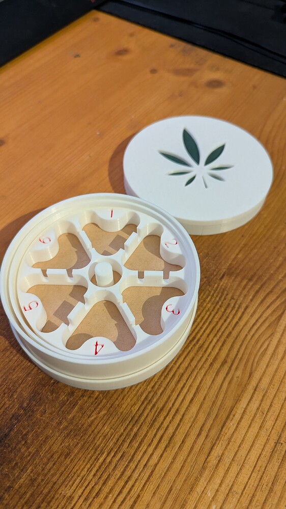 Eine weiße 3D-gedruckte Box mit sechs Fächern zur Samenkeimung, die mit roten Zahlen von 1 bis 6 nummeriert sind. Der Deckel der Box, der daneben liegt, hat einen ausgeschnittenen Marihuana-Blatt-Aufdruck.