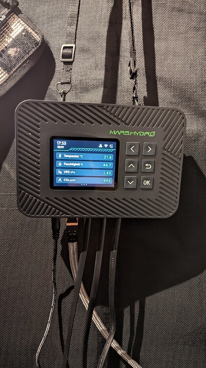 Ein Mars Hydro Controller mit einem Bildschirm, der Temperatur, Luftfeuchtigkeit und CO2-Werte anzeigt, hängt an einem schwarzen Stoffhintergrund.