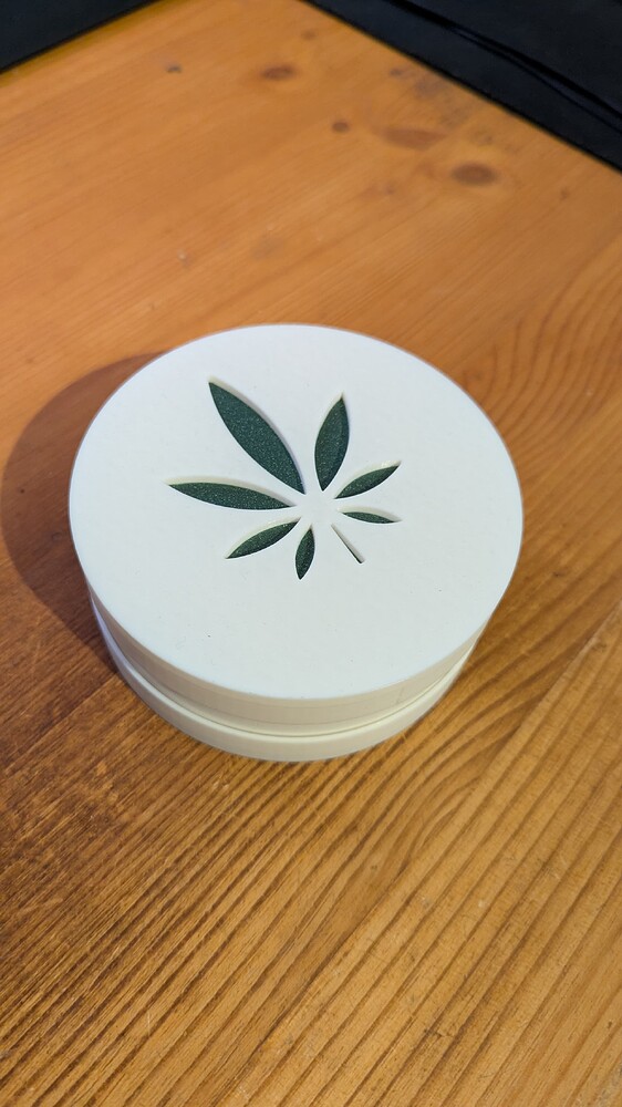 Eine weiße, runde Box mit einem ausgeschnittenen Cannabisblatt-Symbol auf dem Deckel, die auf einer Holzoberfläche steht.